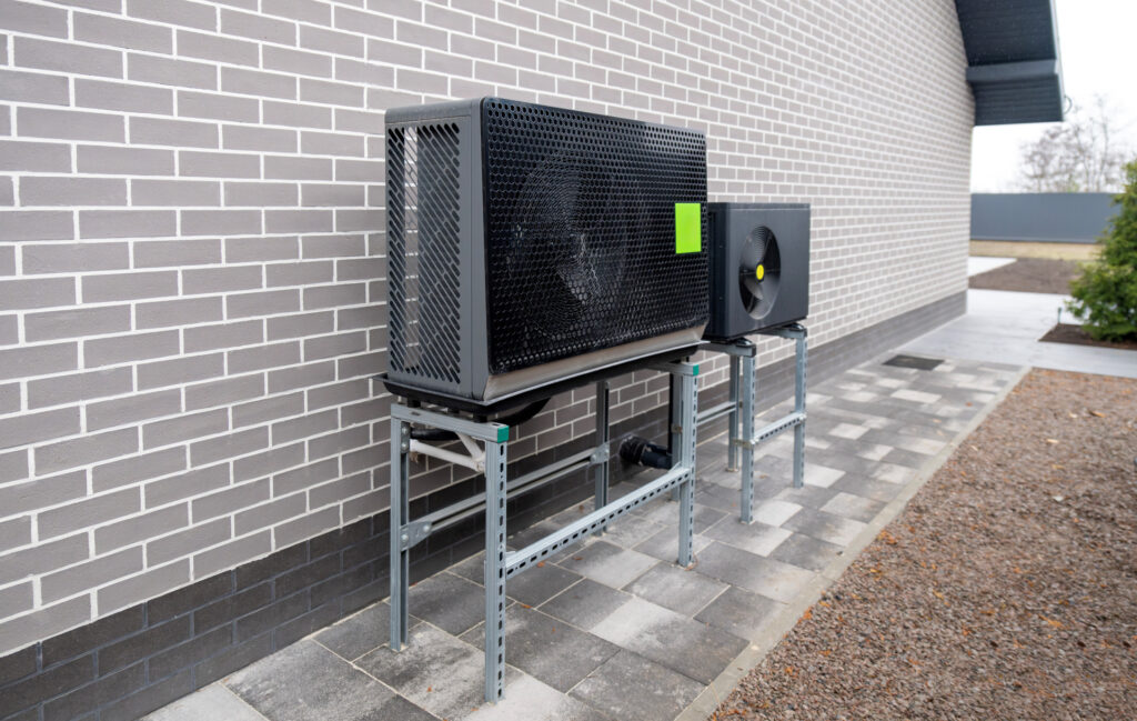 Eine Wärmepumpen-Außeneinheit steht auf Metallständern an einer Hauswand.