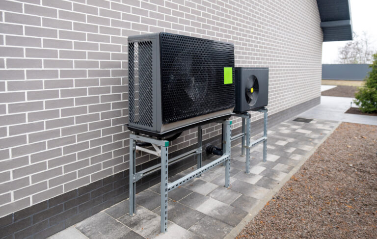 Eine Wärmepumpen-Außeneinheit steht auf Metallständern an einer Hauswand.