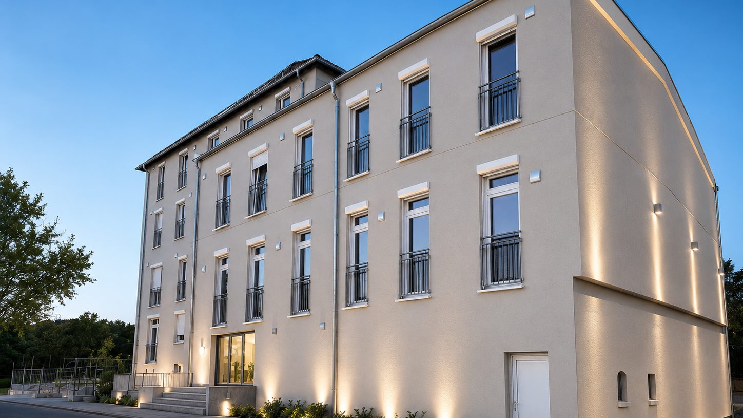 Ein saniertes Mehrfamilienhaus am Abend, von der Straße aus schräg gesehen; die Fassade ist von unten angestrahlt, neben den Fenstern sitzen kleine helle Außenhauben in der Wand.
