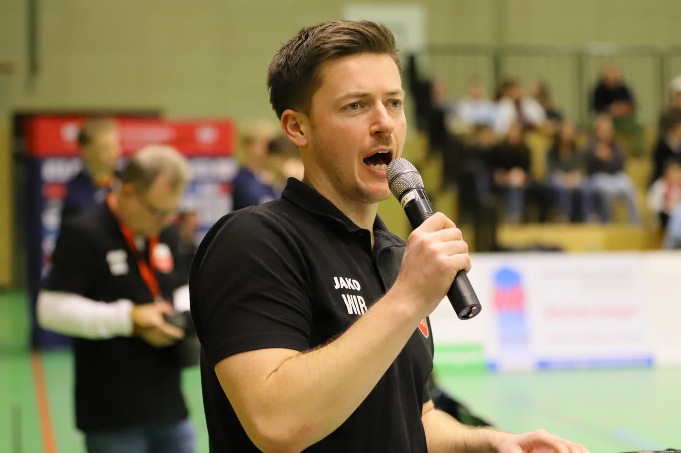 Timo Hensler, Manager des TV Rottenburg, spricht in der Halle mit einem Mikrofon zum Publikum.